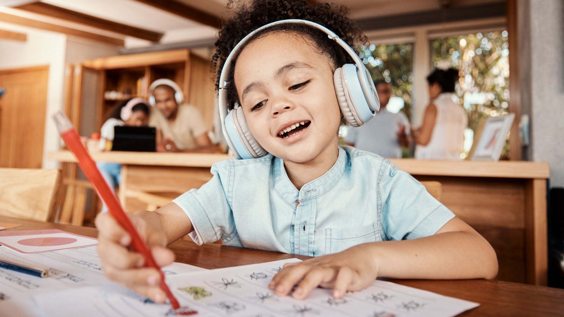 Girl wearing headphones, drawing at a table with family in the background.