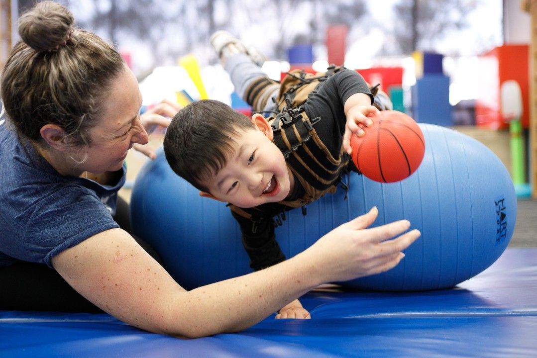 Woman assists child on large blue exercise ball; child smiles, reaching for small ball.