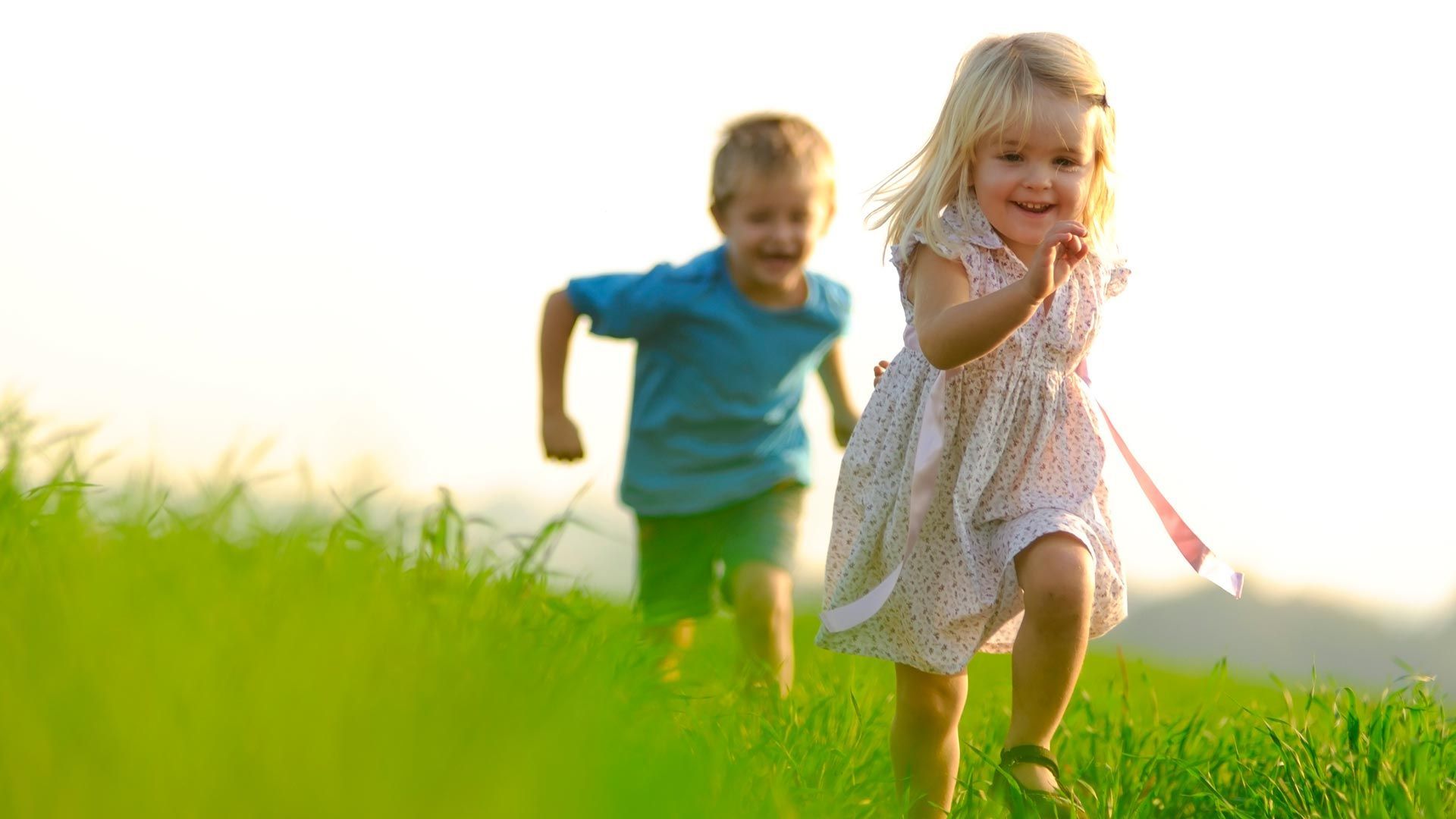 Two children running in a grassy field, smiling. The girl in a dress is closer to the viewer.