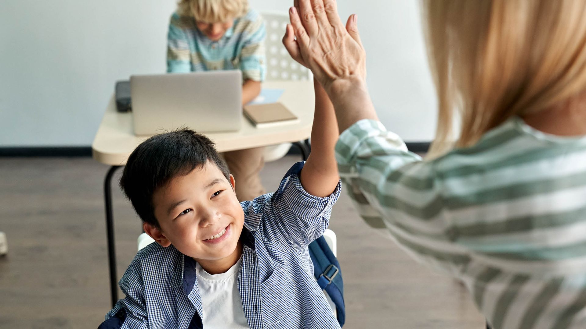 Smiling child gives high-five to a person. Another child works on a laptop at a desk in the background.