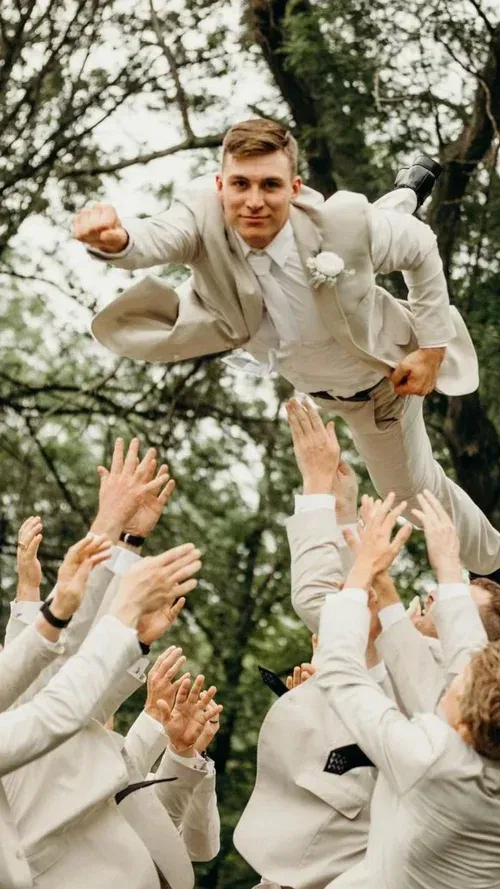 Groom in beige suit being tossed into the air by groomsmen, outdoors.