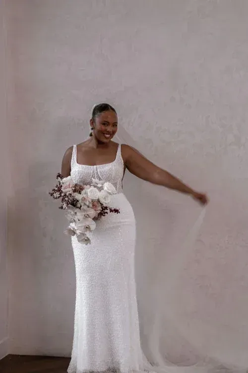 Bride in a white gown holding a bouquet, arm outstretched, veil trailing, smiling.