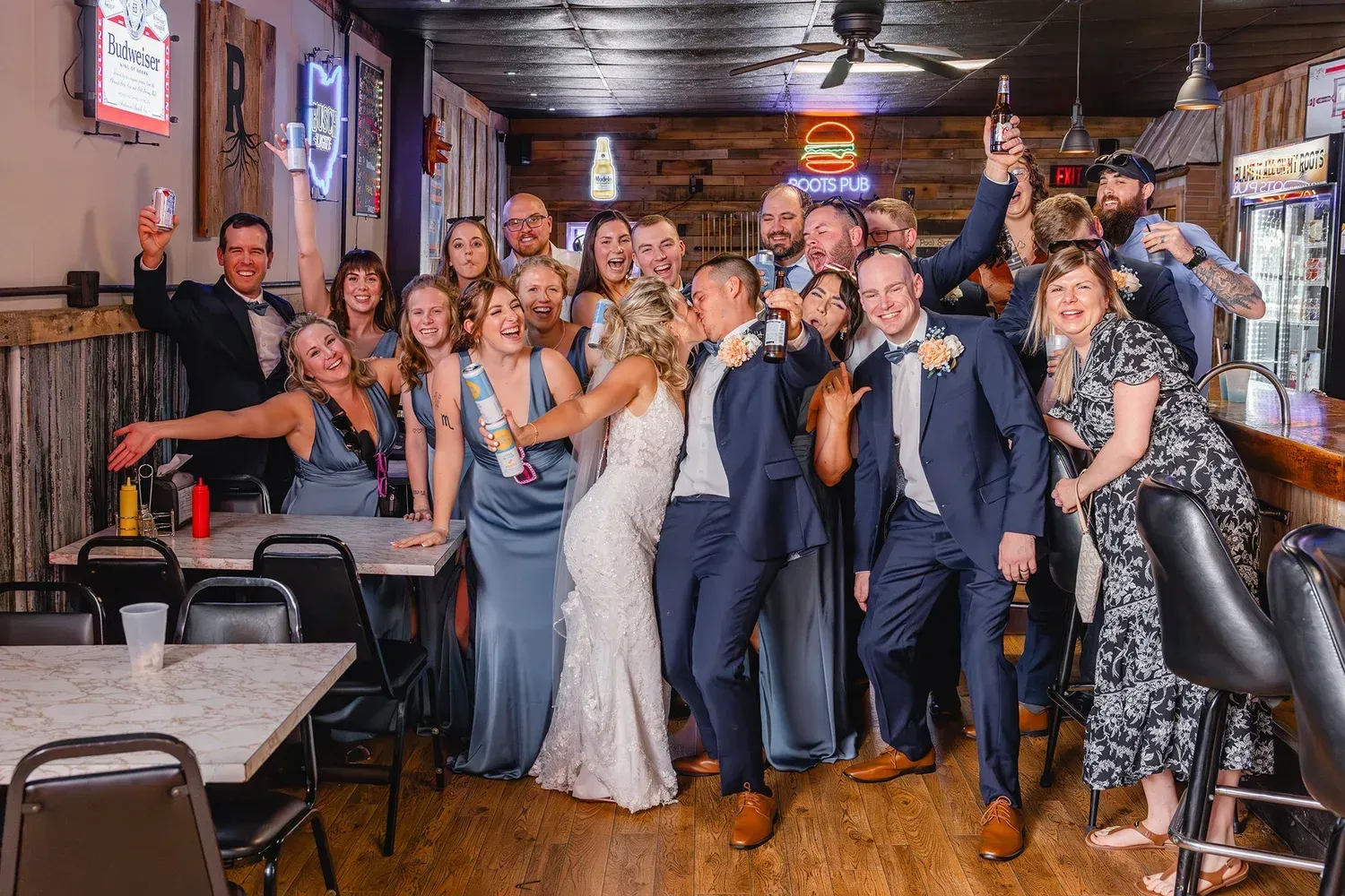 Wedding party cheers in a rustic bar. Bride and groom surrounded by smiling friends, raising drinks.