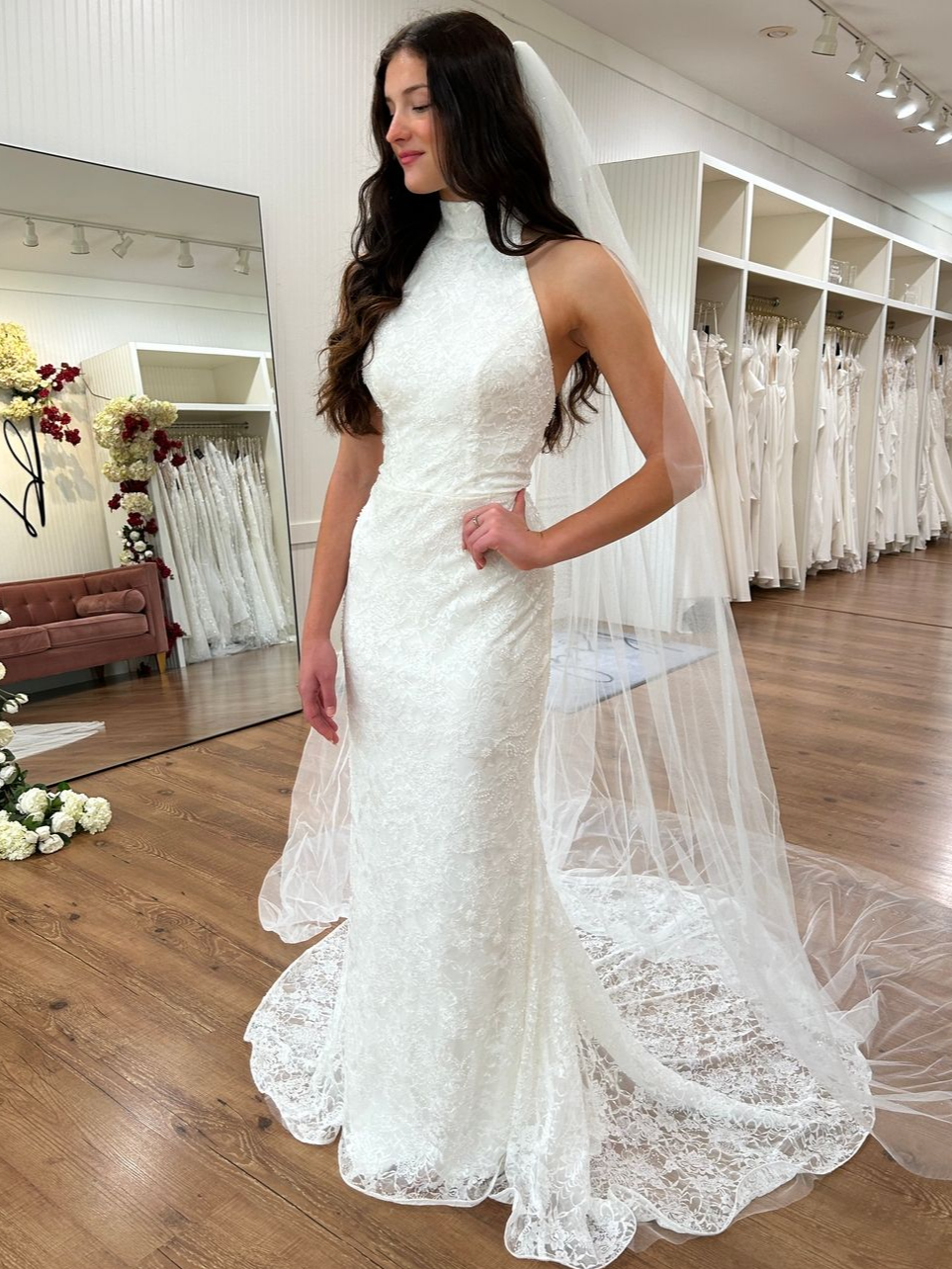 Bride in a white lace halter wedding dress with a veil, standing in a bridal shop.