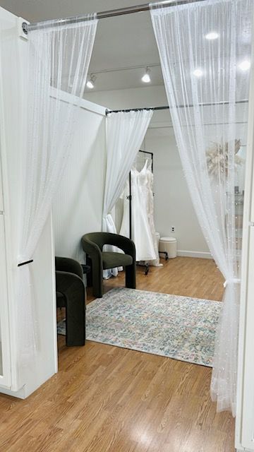 A bridal boutique with a viewing area. Two white curtains frame a room with a full-length mirror, dresses, and chairs