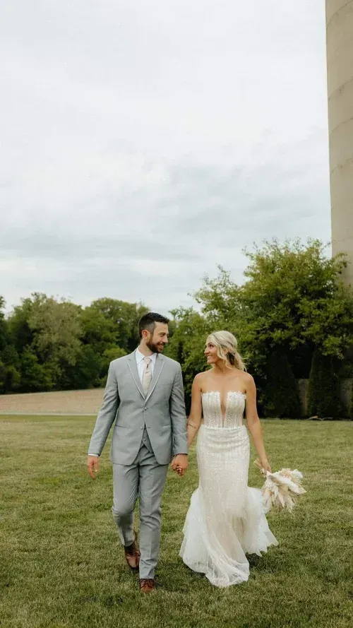 A bride and groom holding hands, smiling, walking on grass, wearing wedding attire. Cloudy sky, tower in background.