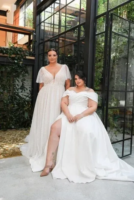 Two women in wedding dresses, one with a leg out, standing in front of a glass wall.