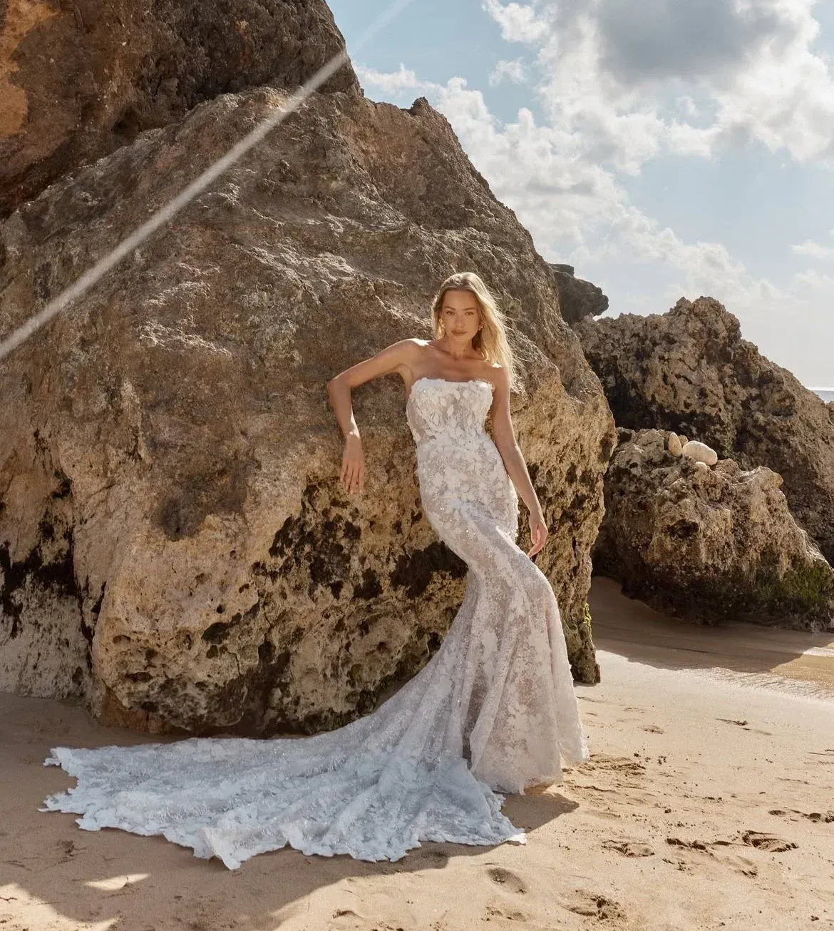 Woman in a strapless lace wedding dress leans against a large rock on a sunny beach.