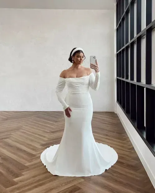 Woman in white off-the-shoulder wedding dress with long sleeves poses near window, taking a selfie.