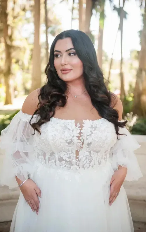 Woman in a white off-the-shoulder wedding dress, hands on hips, looks to the side, outdoor setting.