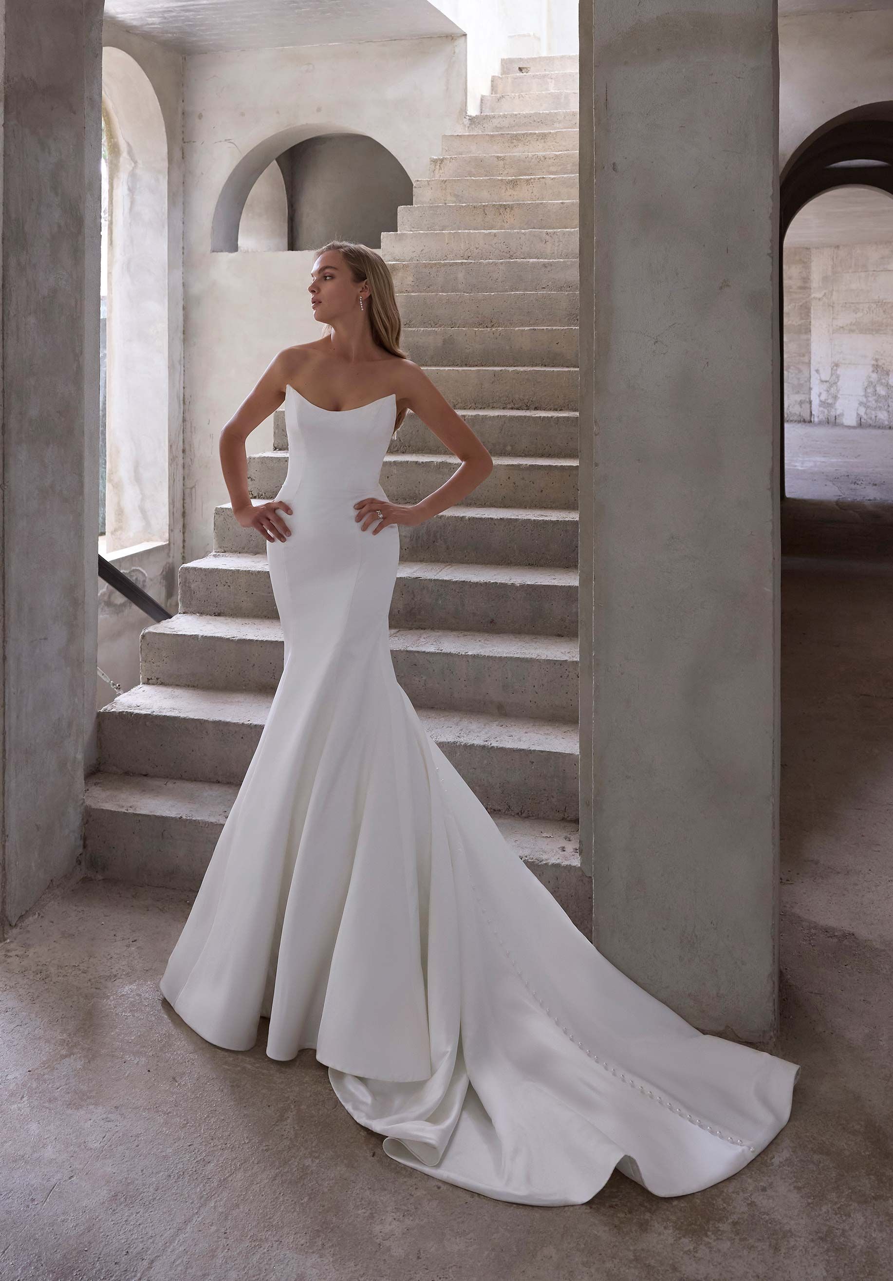 Bride in white strapless gown stands on stairs, hands on hips. Interior with arches, stone.