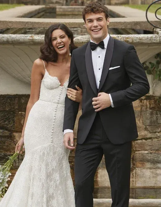 Smiling bride and groom in formal attire, posing outdoors. Bride in white gown, groom in black tux.