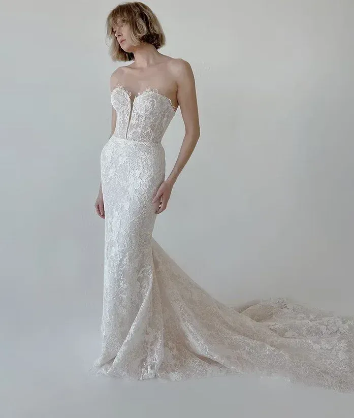 Woman in strapless, fitted, lace wedding gown with a train, posing in front of a white backdrop.