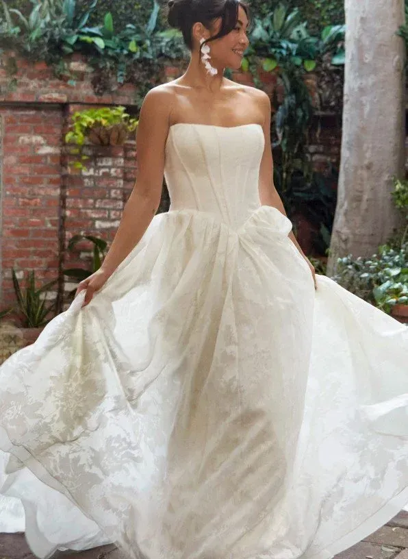 Woman in a strapless white wedding dress, smiling, holding the dress in a garden setting.