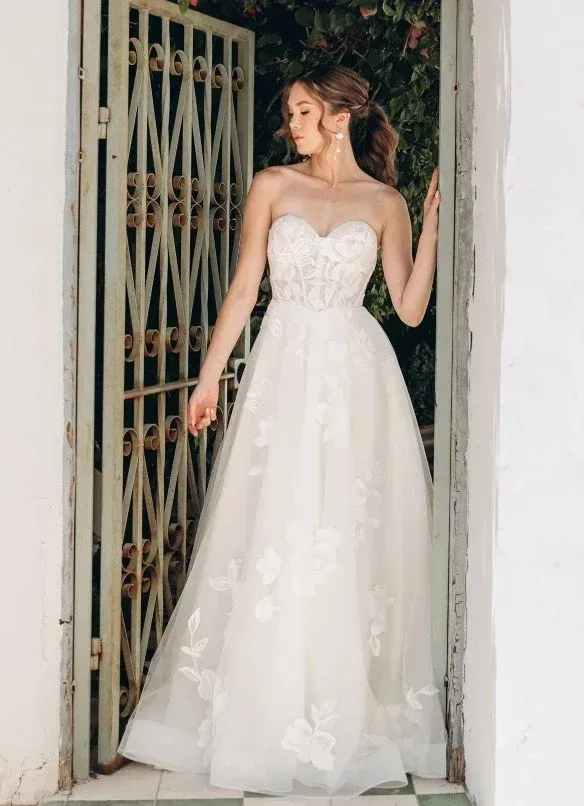 Woman in strapless white wedding dress stands in doorway, holding gate.