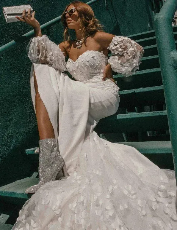 Woman in a white gown with feathered train, sparkly boots, and sunglasses poses on teal steps, holding a small purse.