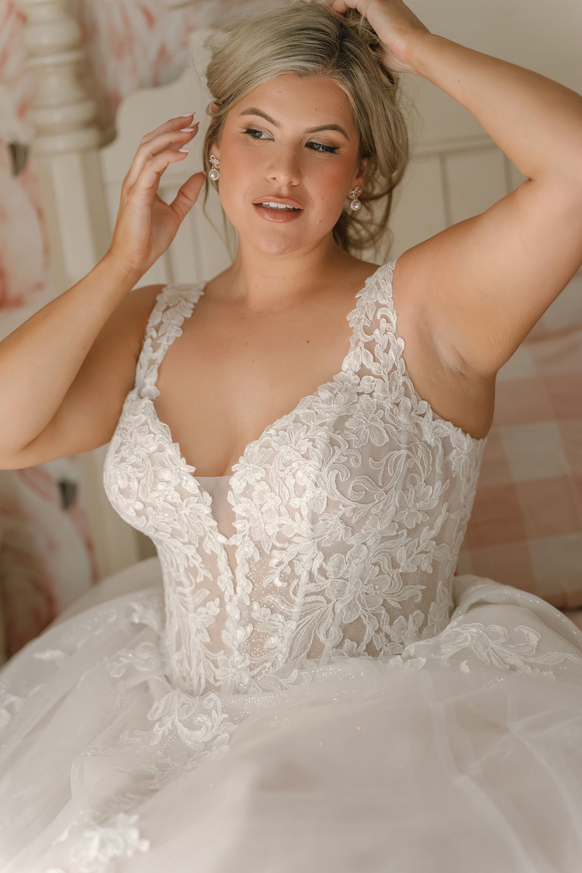 Bride in a white lace wedding dress, adjusting her hair in a softly lit room.