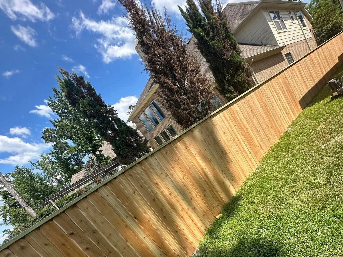 Tilted view of a wooden backyard fence beside green grass, tall trees, and a house under a blue sky