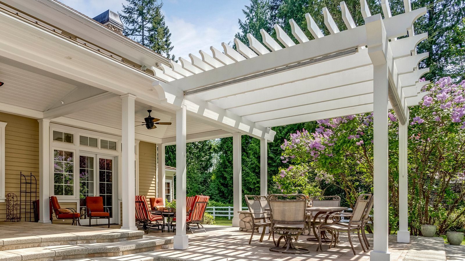 Patio with white pergola, outdoor dining table, red chairs, and greenery beside a house