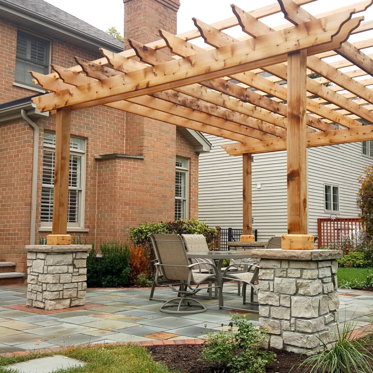 Backyard patio with a wooden pergola, stone columns, and outdoor chairs beside a brick house
