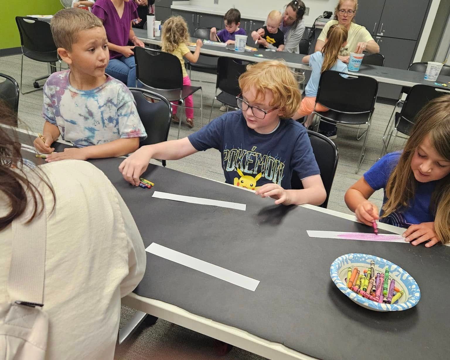 Children at a table crafting, others in background.  Black table with white strips, crayons in a bowl.