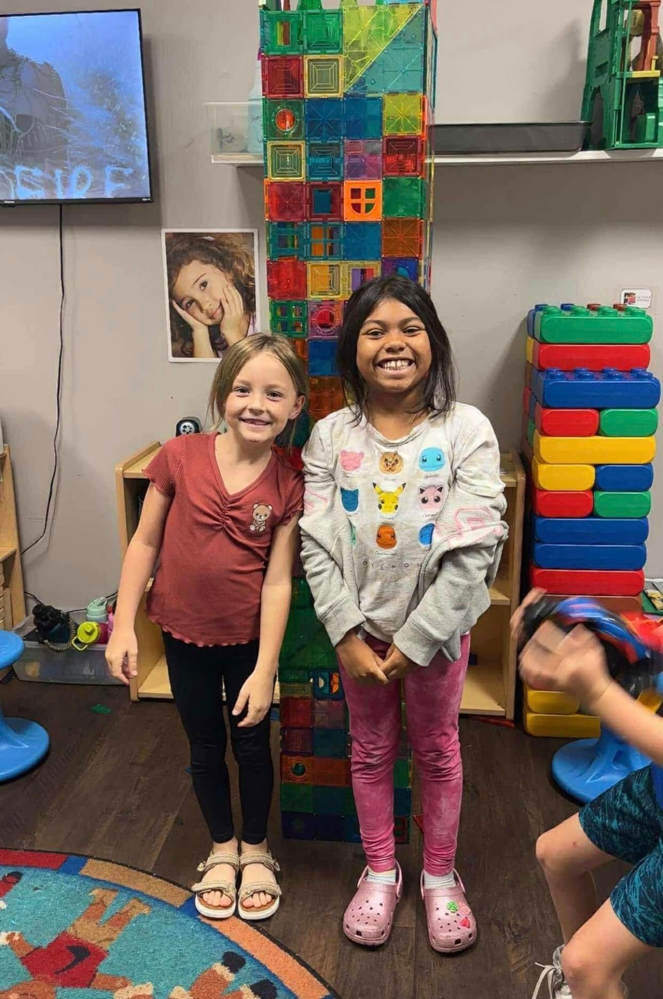 Two children smiling next to a tall, colorful tower made of building blocks in a classroom.