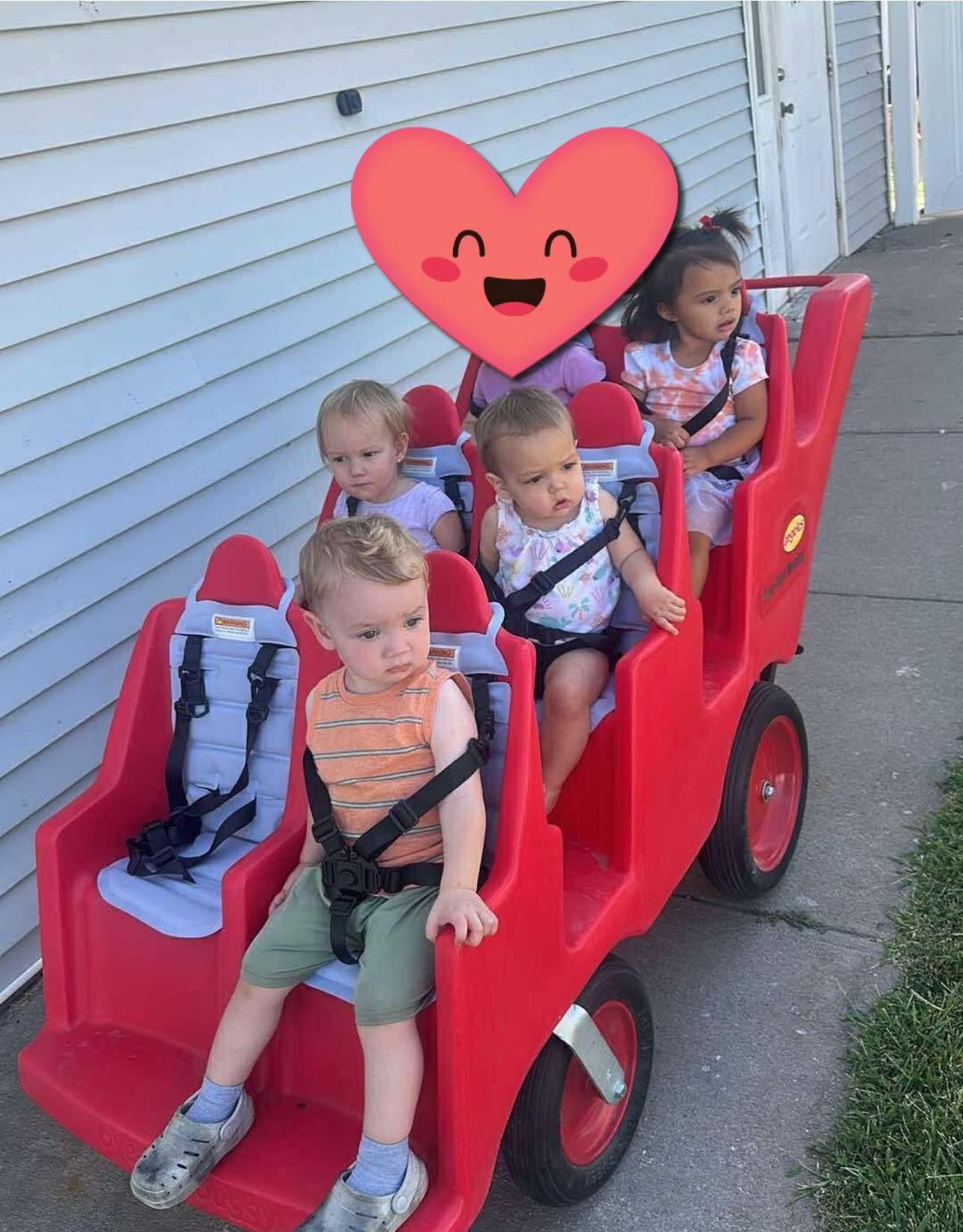 Children in red stroller-like wagon with safety harnesses. Heart emoji superimposed on top. Outdoor setting.