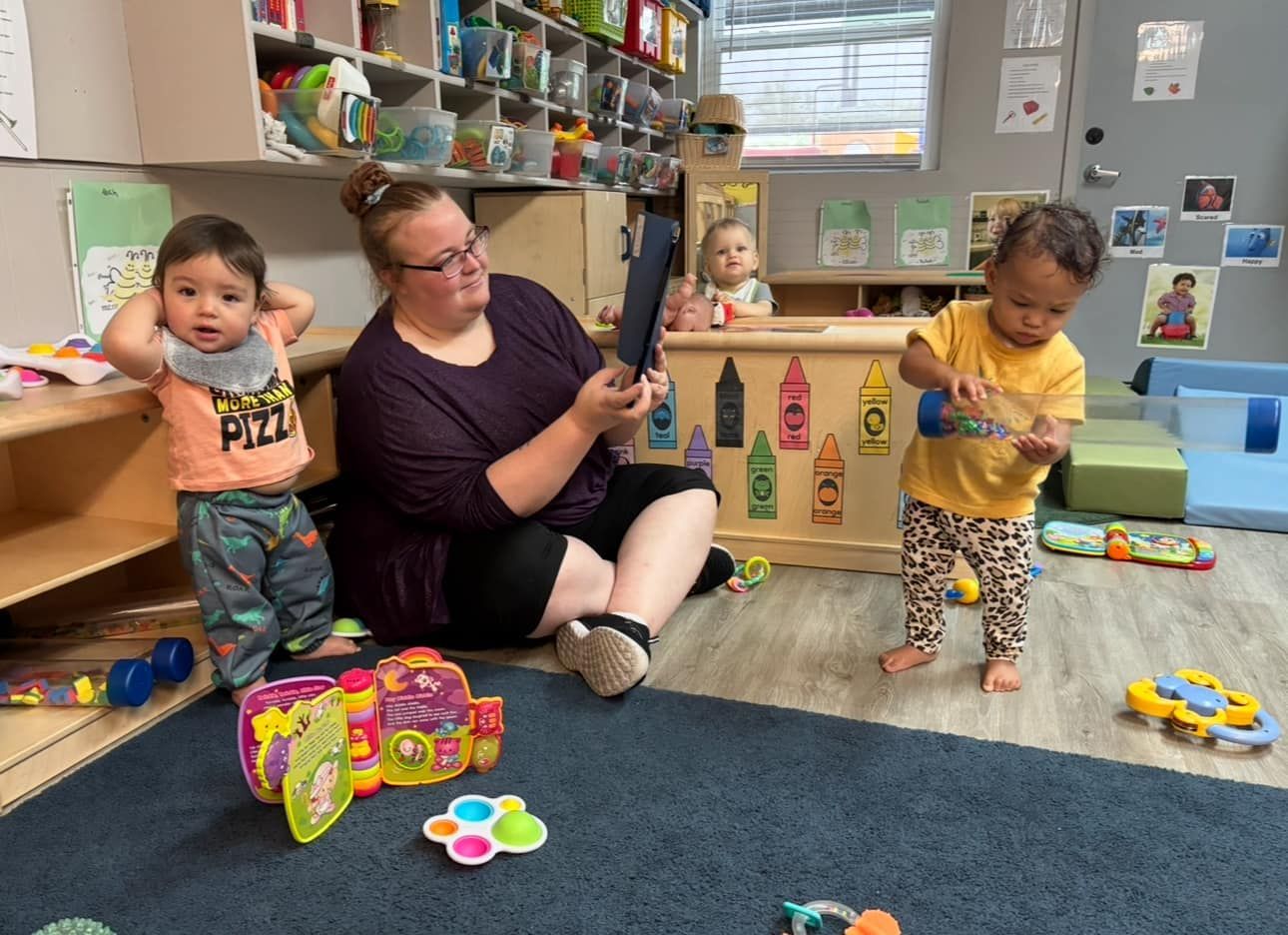A childcare provider reads a book to children on a classroom floor. Toys and shelves are visible.