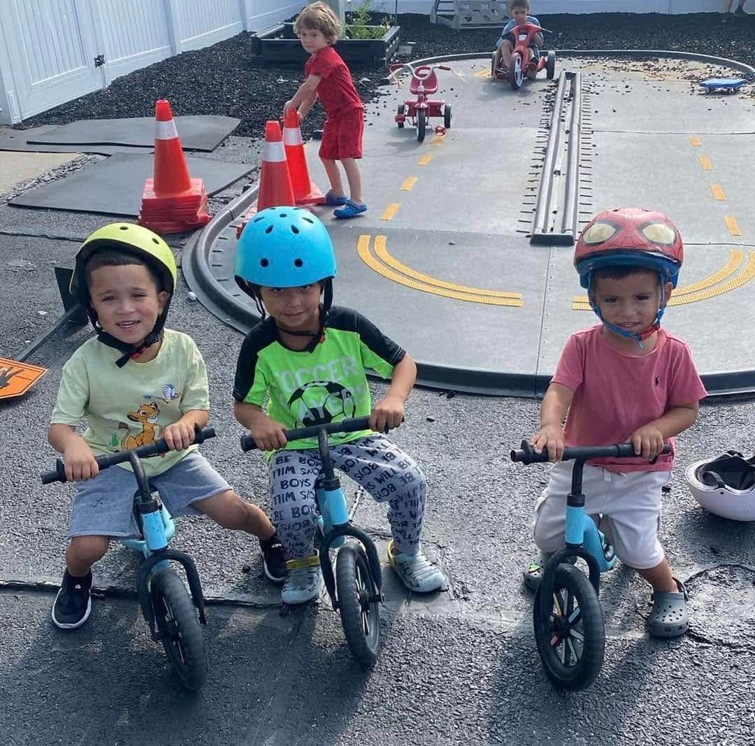 Three children on balance bikes with helmets, another child plays in the background near a road track with cones.