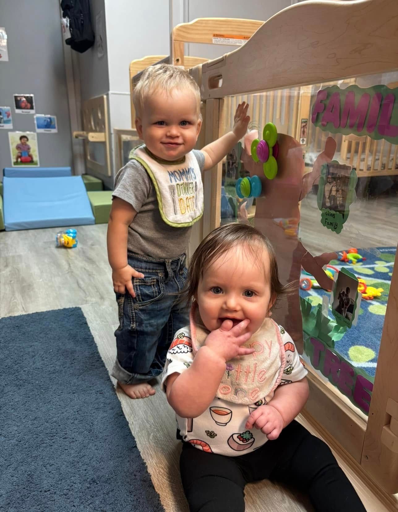Two babies in a playroom: one stands by glass, other sits with finger in mouth, both smiling.