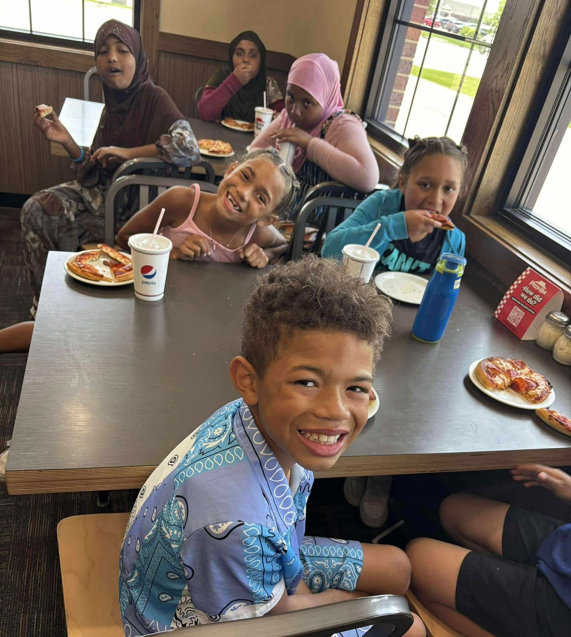 Group of kids smiling around a table with pizza and drinks at a restaurant.