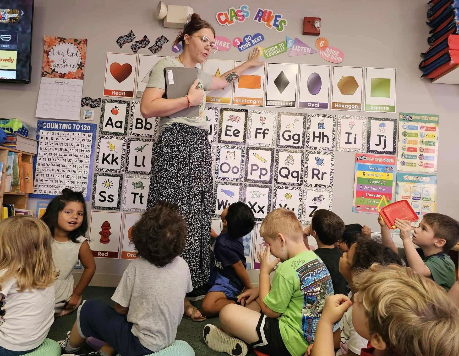 Teacher pointing at letters on a wall, surrounded by children sitting on the floor in a classroom.