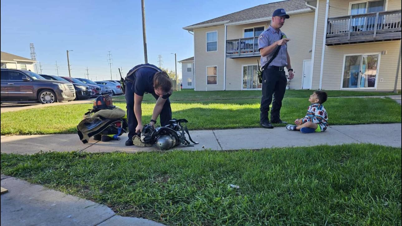 A firefighter tends to equipment on a sidewalk while a child sits nearby. Another person stands close.