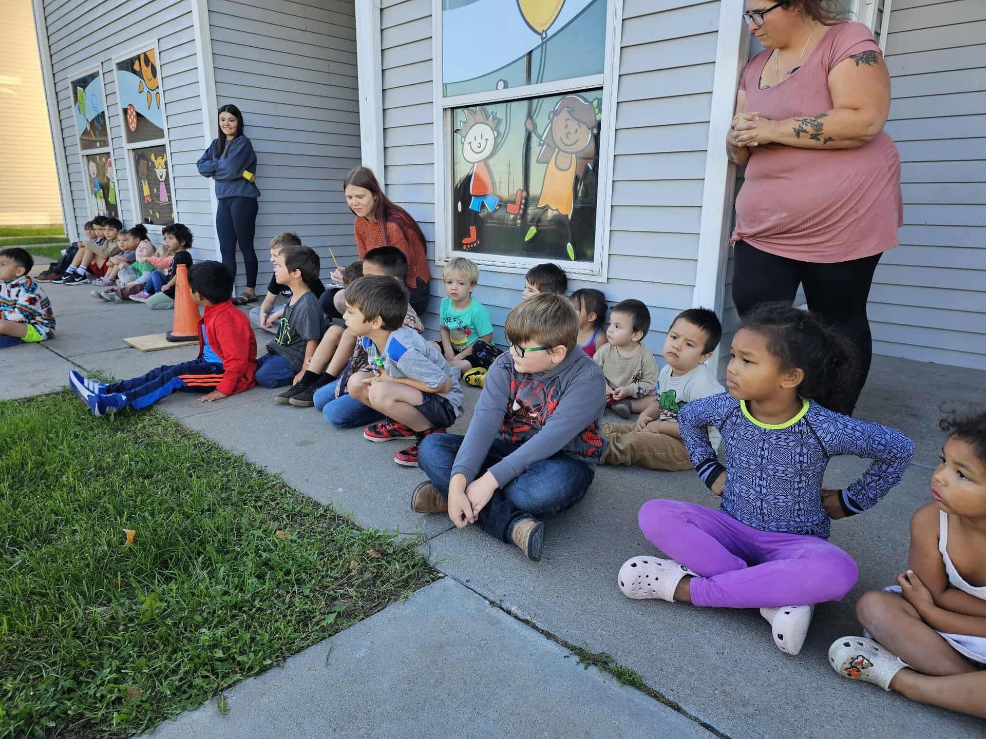 Children seated outside a building with adults watching.