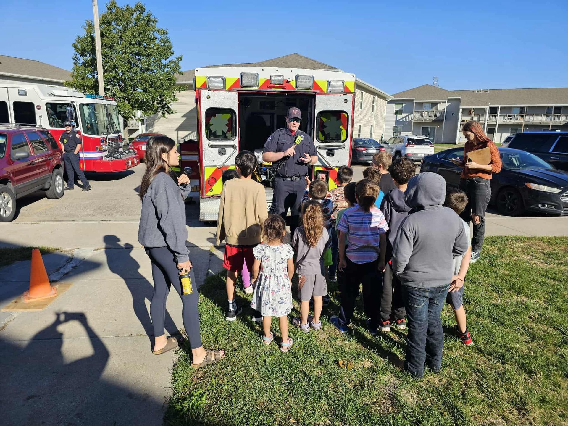 Children listen to a firefighter in front of an ambulance; a fire truck is visible nearby.