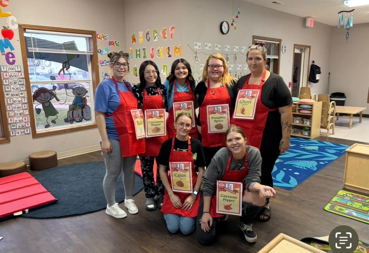 Group of people wearing red aprons in a classroom. They are smiling and holding recipe cards.