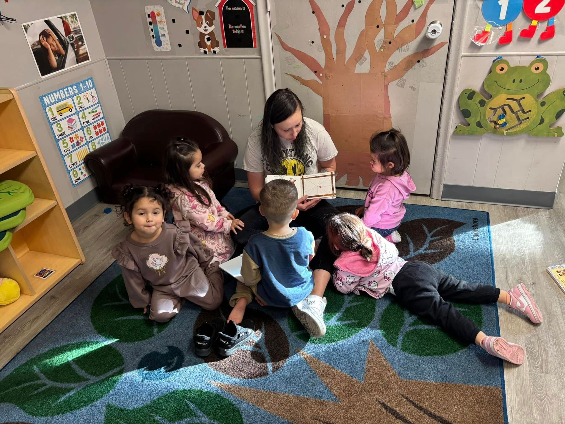 Teacher reading to children on a leaf-themed rug in a classroom.