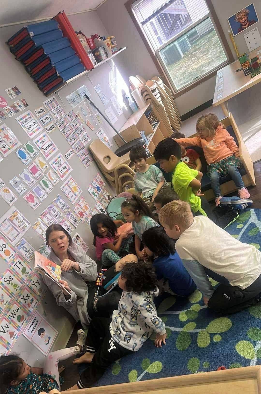 Children gathered around a person reading a book in a classroom with charts on the wall.