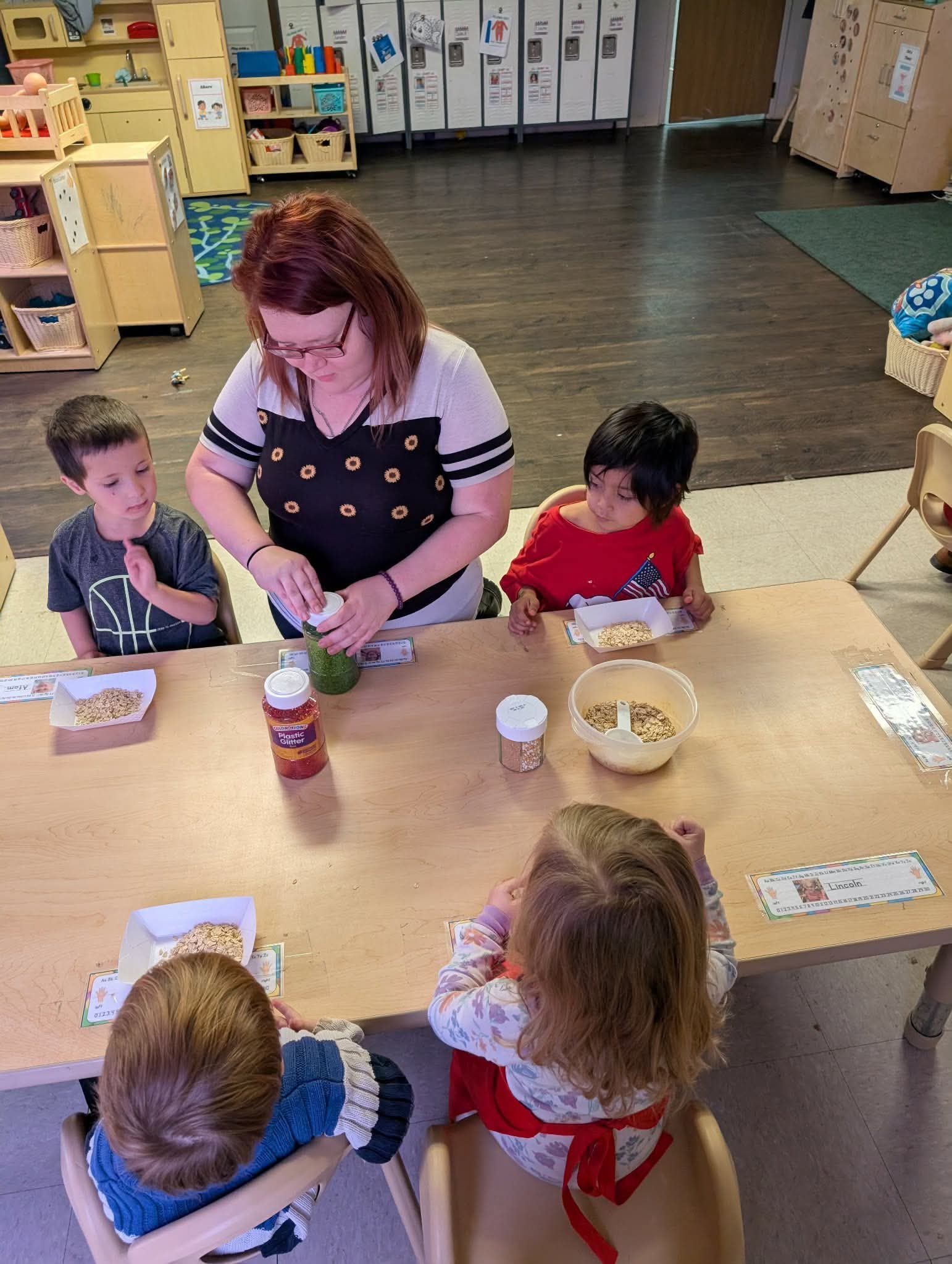 A teacher prepares food at a table for children in a brightly lit classroom.