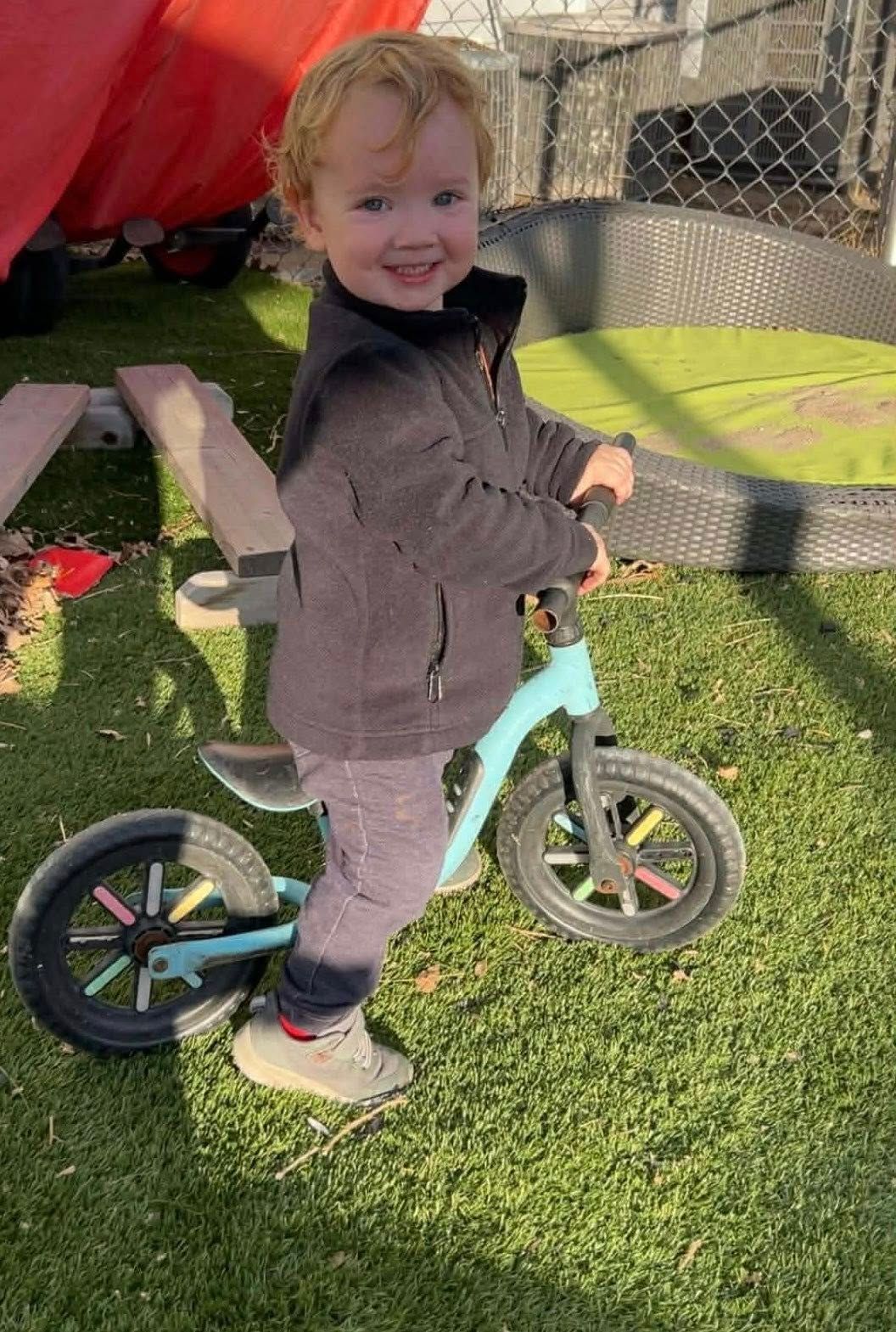 Young child smiling on a blue balance bike, standing on green grass.