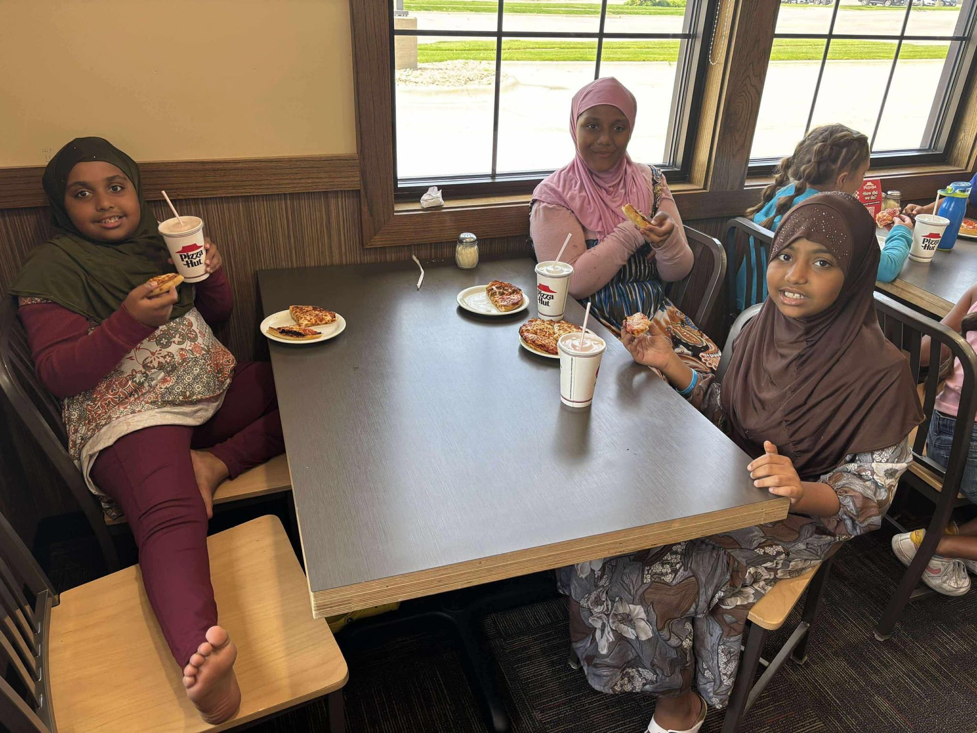 Three people in headscarves at a table, eating and drinking. Two are smiling, one looks at the camera. Inside a restaurant.