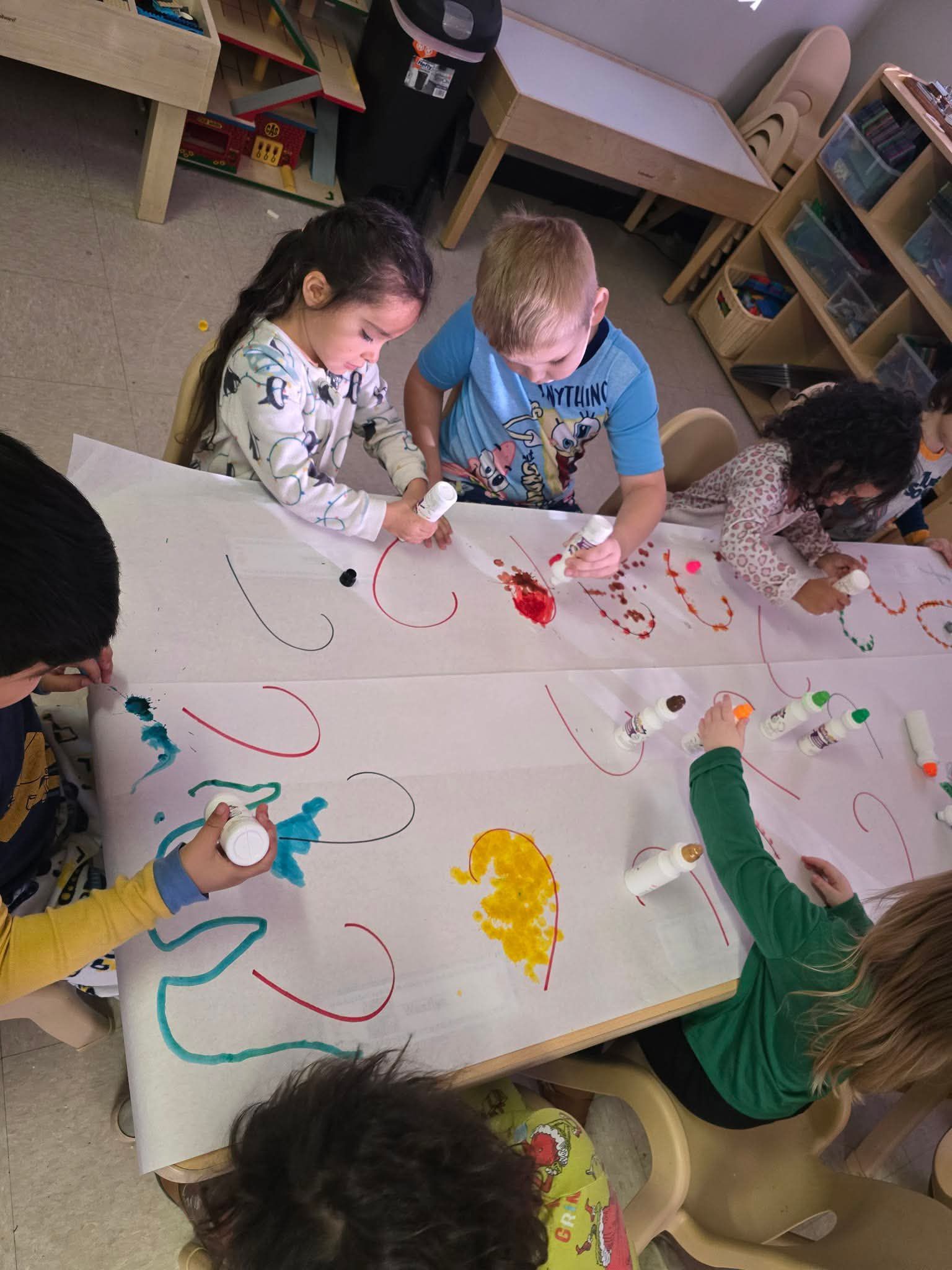 Children painting on a large sheet of paper on a table, using paint tubes of various colors in a classroom.