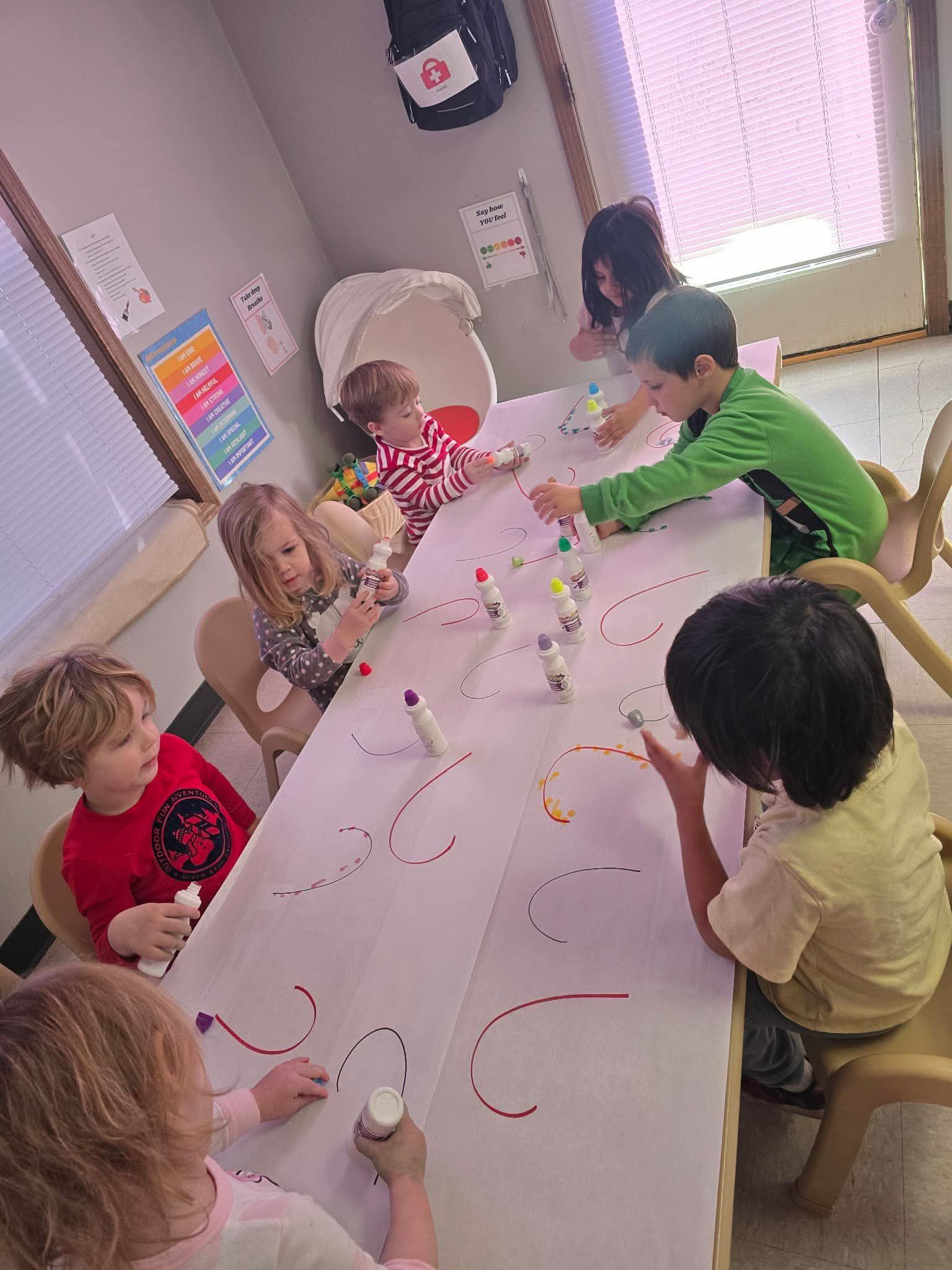 Children sit around a large table, coloring with markers. A window and colorful posters are visible.
