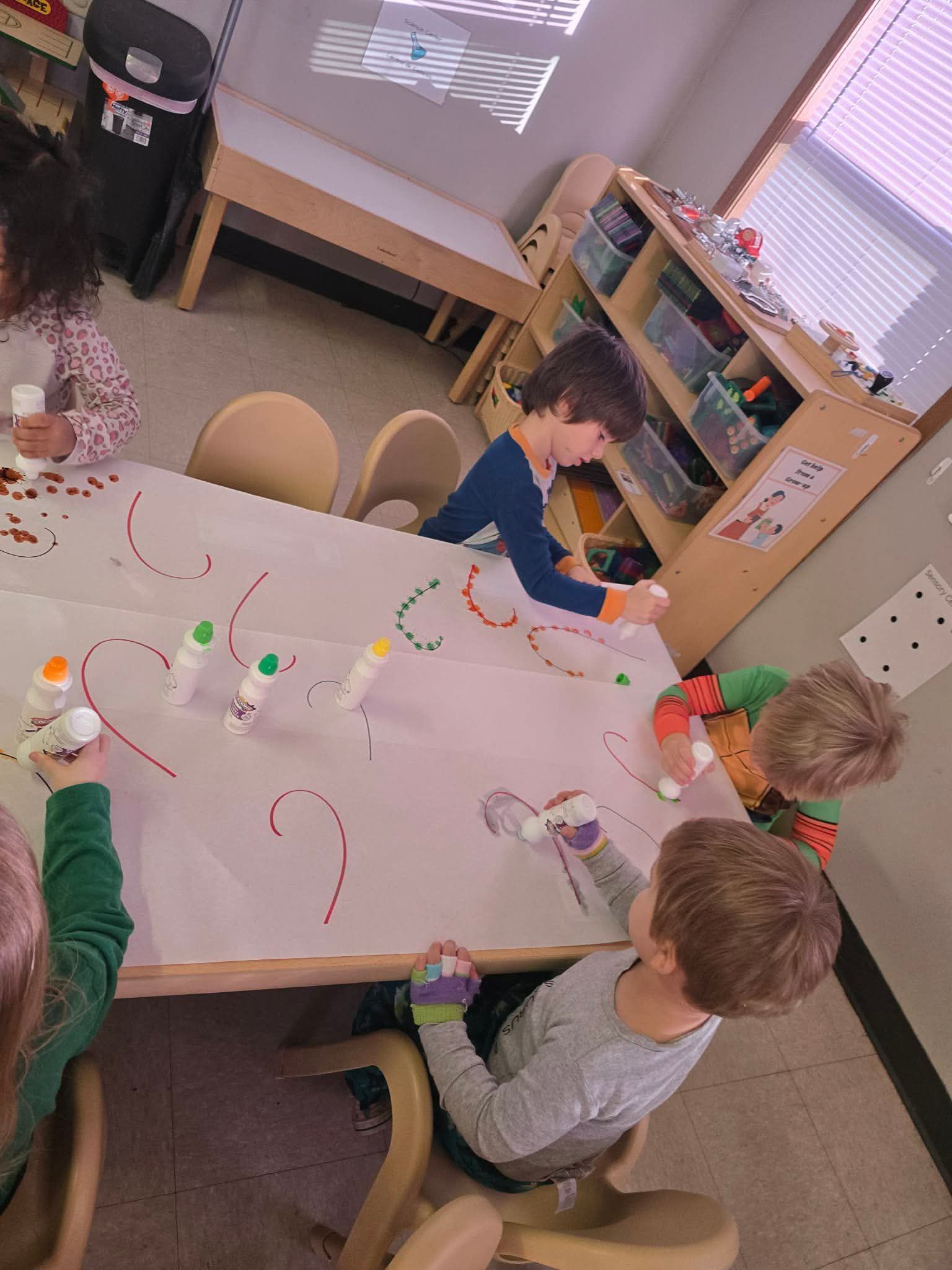 Children painting on a large sheet of paper at a table in a classroom.