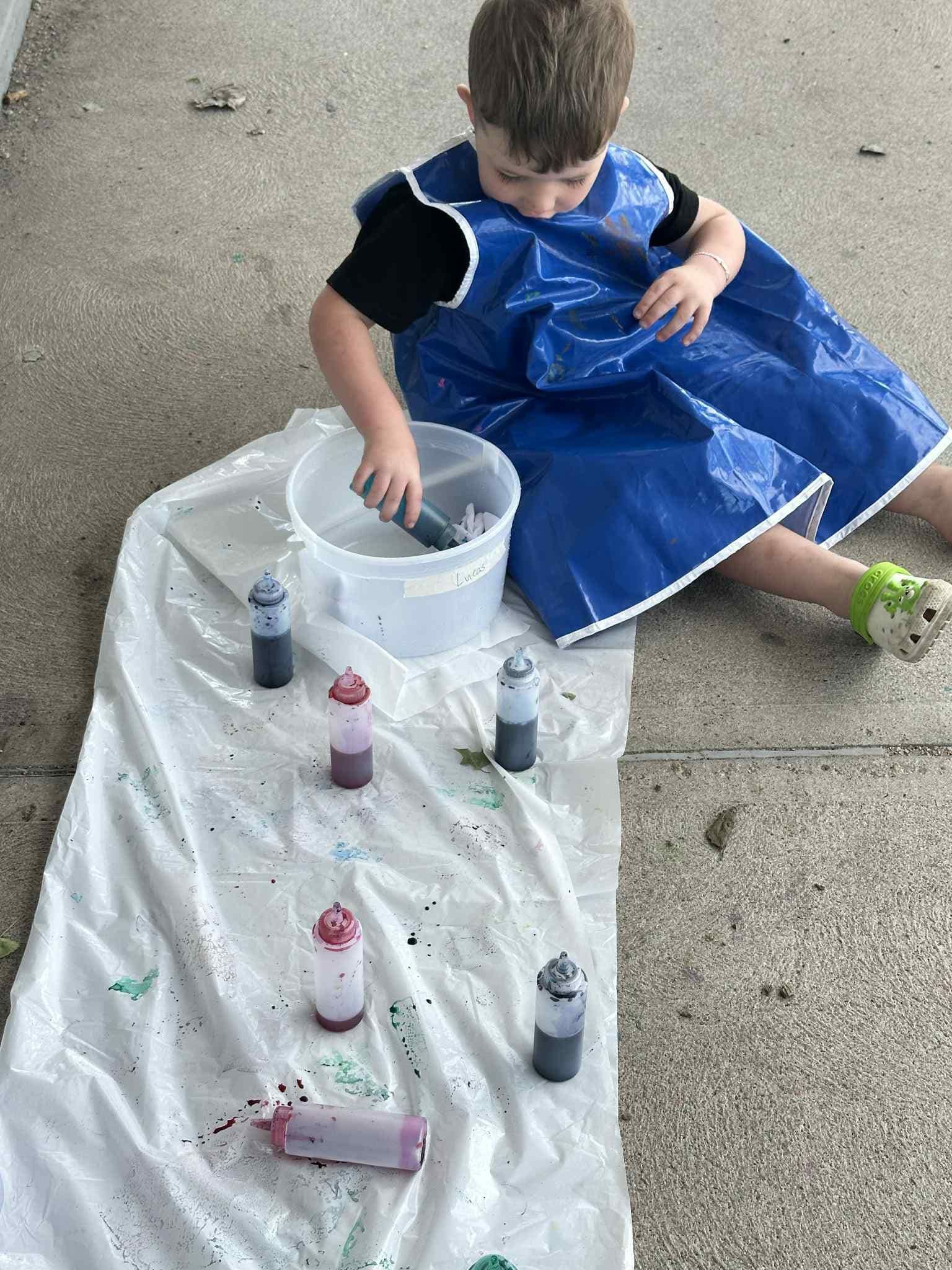 Child tie-dyeing fabric outside, wearing blue apron. Dyes and bucket on a plastic sheet, concrete ground.