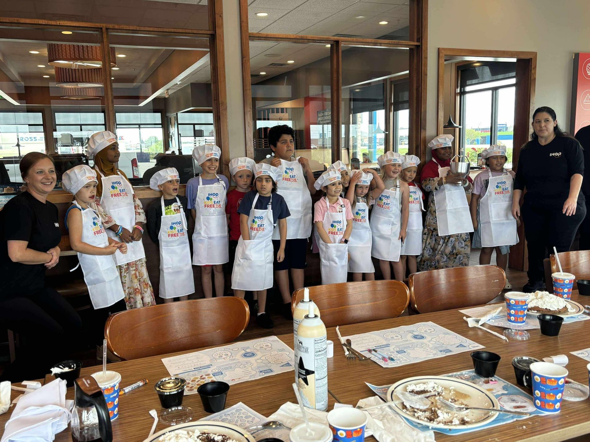 Children in aprons and chef hats pose with adults at a table with food, possibly a cooking class in a restaurant.