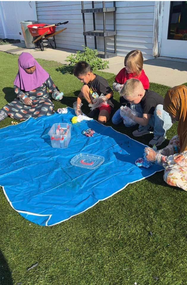 Children gathered outdoors around a blue tarp, playing with items from plastic containers in the grass.