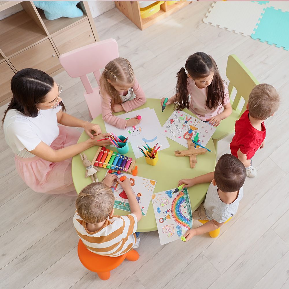 Teacher and children drawing at a round table in a brightly lit room. Crayons, art supplies, and finished artwork present.