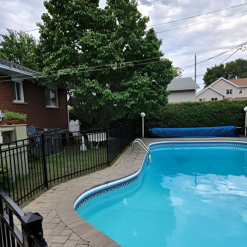 Piscine à l'arrière d'un jardin entouré d'une clôture noire et d'une terrasse en briques. Une piscine couverte se trouve devant des arbres verdoyants et des maisons, sous un ciel nuageux.