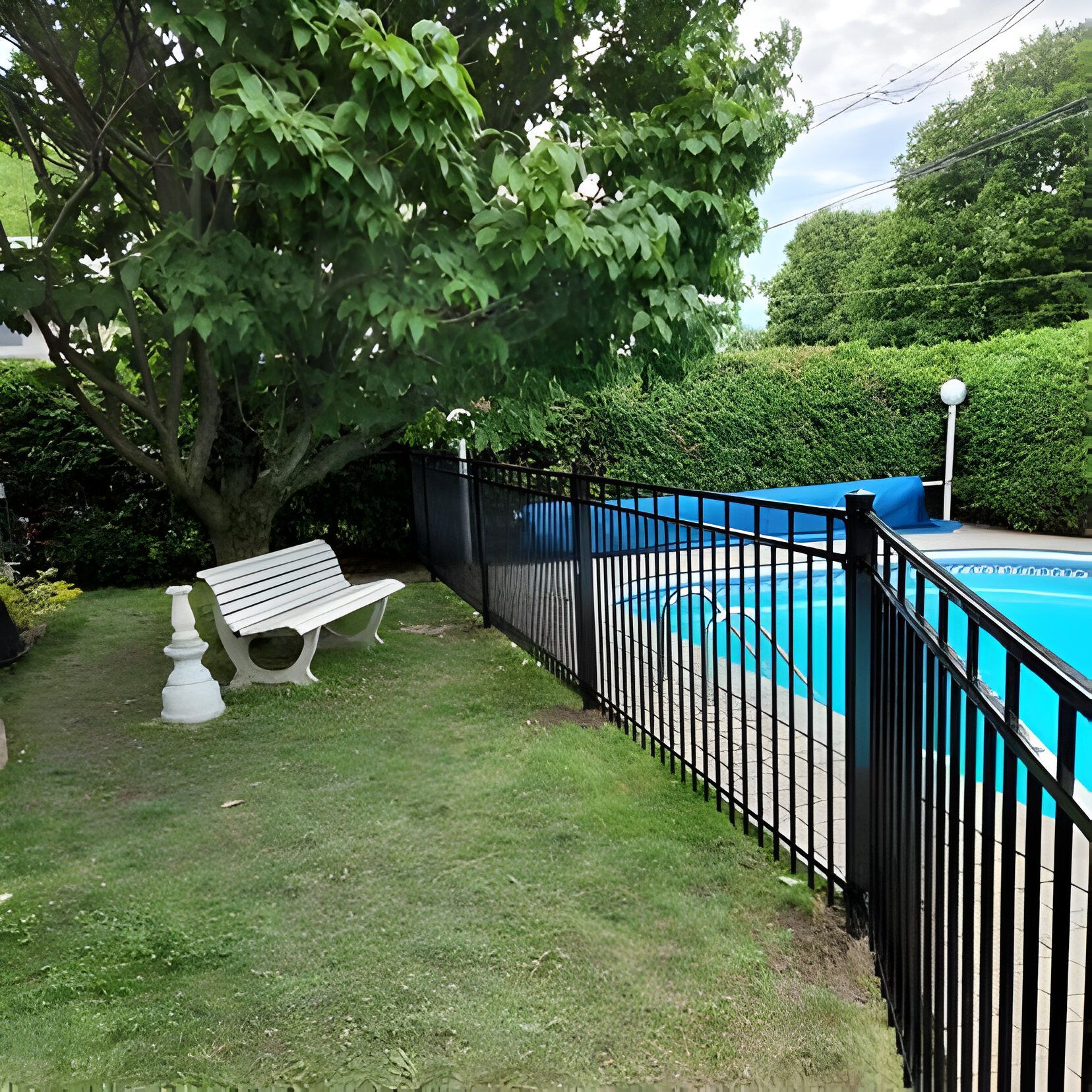 Une piscine entourée d'une clôture noire, avec un banc blanc sur la pelouse, un arbre et une haie, par une journée partiellement nuageuse.